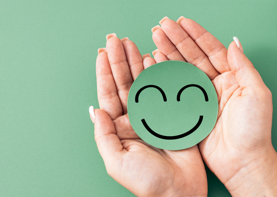 Hands holding a cardboard cutout smiley face on green background, satisfaction, well being, happiness, mental health,